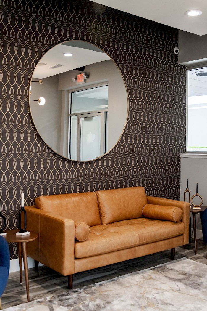 A tan leather sofa under a round gold mirror on the patterned feature wall of the lounge